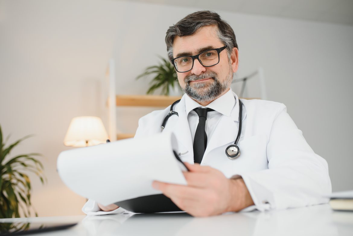 Portrait of friendly smiling senior adult healthcare professional therapist sitting at workplace. Happy confident older male doctor physician wearing white medical coat stethoscope looking at camera