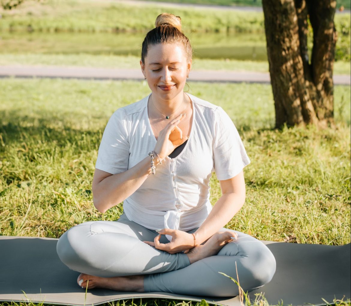 Yoga and meditation, mind health and mental harmony concept. Calm smiling young yogi woman sitting in lotus position on sports mat, practicing meditation in park at sunrise. Active healthy lifestyle